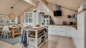 Moderni keittiö, jossa on puiset kaapit, ruokailutila pöytineen ja tuoleineen sekä suuri ikkuna, josta tulee runsaasti luonnonvaloa.