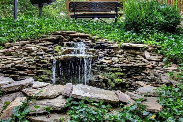 A stone waterfall surrounded by rocks and greenery, with a bench in the background. - PERFECT LOCATION-Dollywood-Spa-Shuffleboard-Hot Tub-Swing-Wooded-Custom Decor (Pigeon Forge)