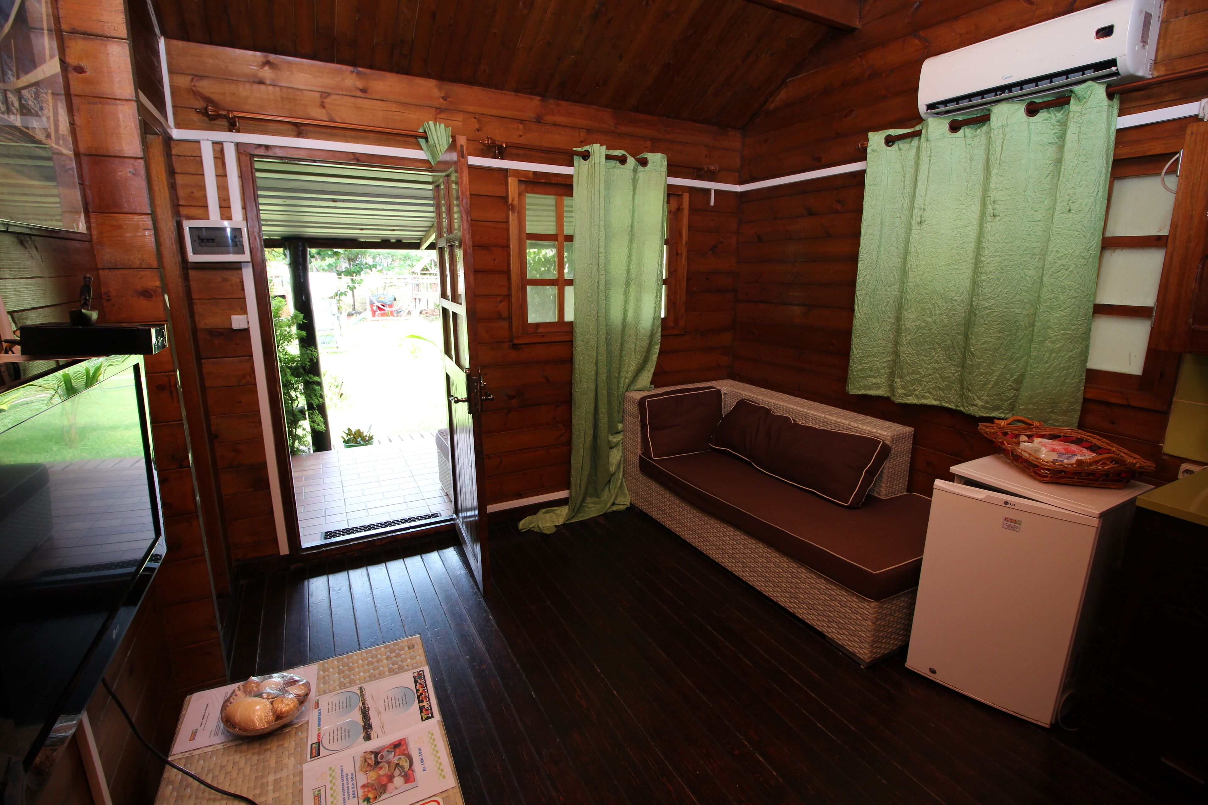 A wooden cabin interior with a wooden floor, a brown sofa, a small refrigerator, and a window with a green curtain.