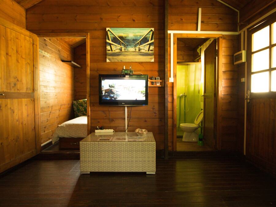 Interior de una cabaña de madera con televisión sobre un mueble bajo, una cama, una mesita y un baño visible a través de una puerta abierta.
