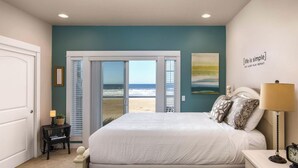 A bedroom with a large bed, a nightstand, a lamp, a clock, a framed picture, and a view of the beach.