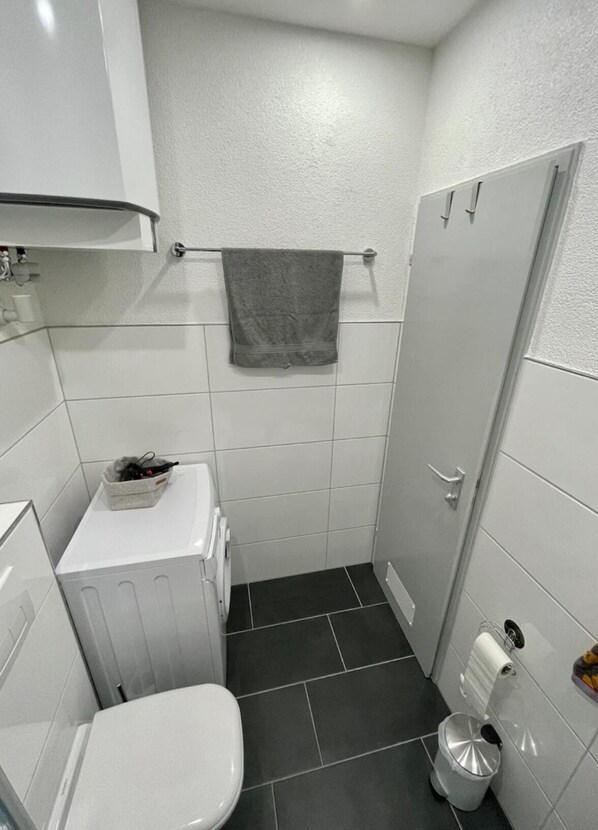 Ett litet, modernt badrum med vita kakelplattor, en vit toalett, en handduksställning med ett handduk, en liten papperskorg och ett vitt skåp.