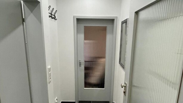 Un couloir étroit, doté d’un sol carrelé foncé, de murs blancs et d’une porte avec une poignée en métal.