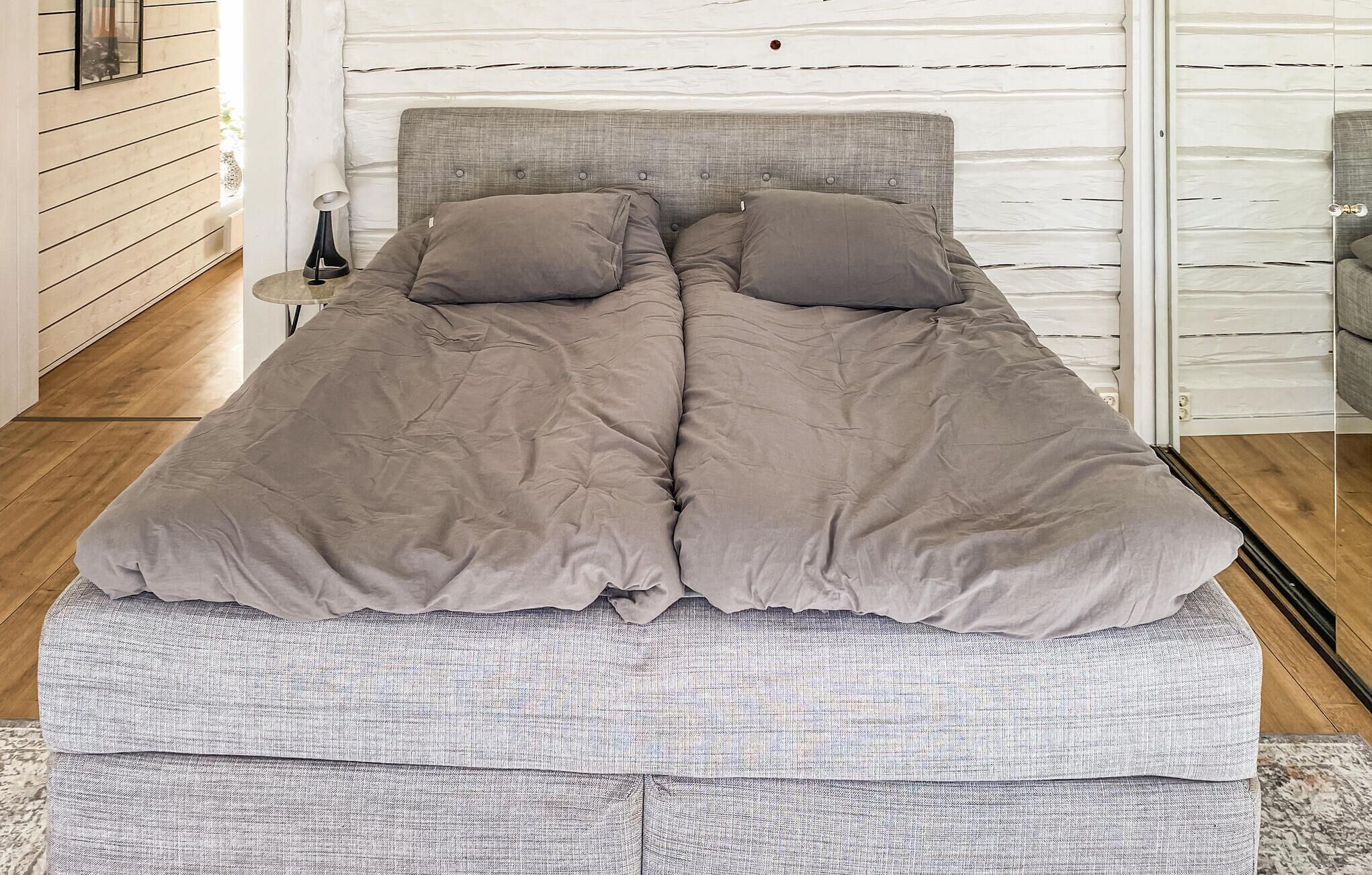 Un lit avec une tête de lit grise et deux oreillers, un banc gris et un mur blanc orné de lignes horizontales.