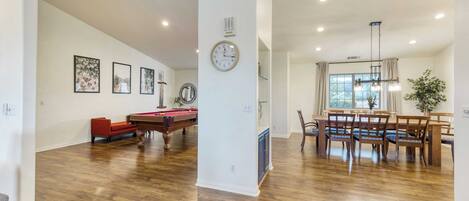 A spacious living room with a pool table, red sofa, and a dining area with wooden chairs and a chandelier.