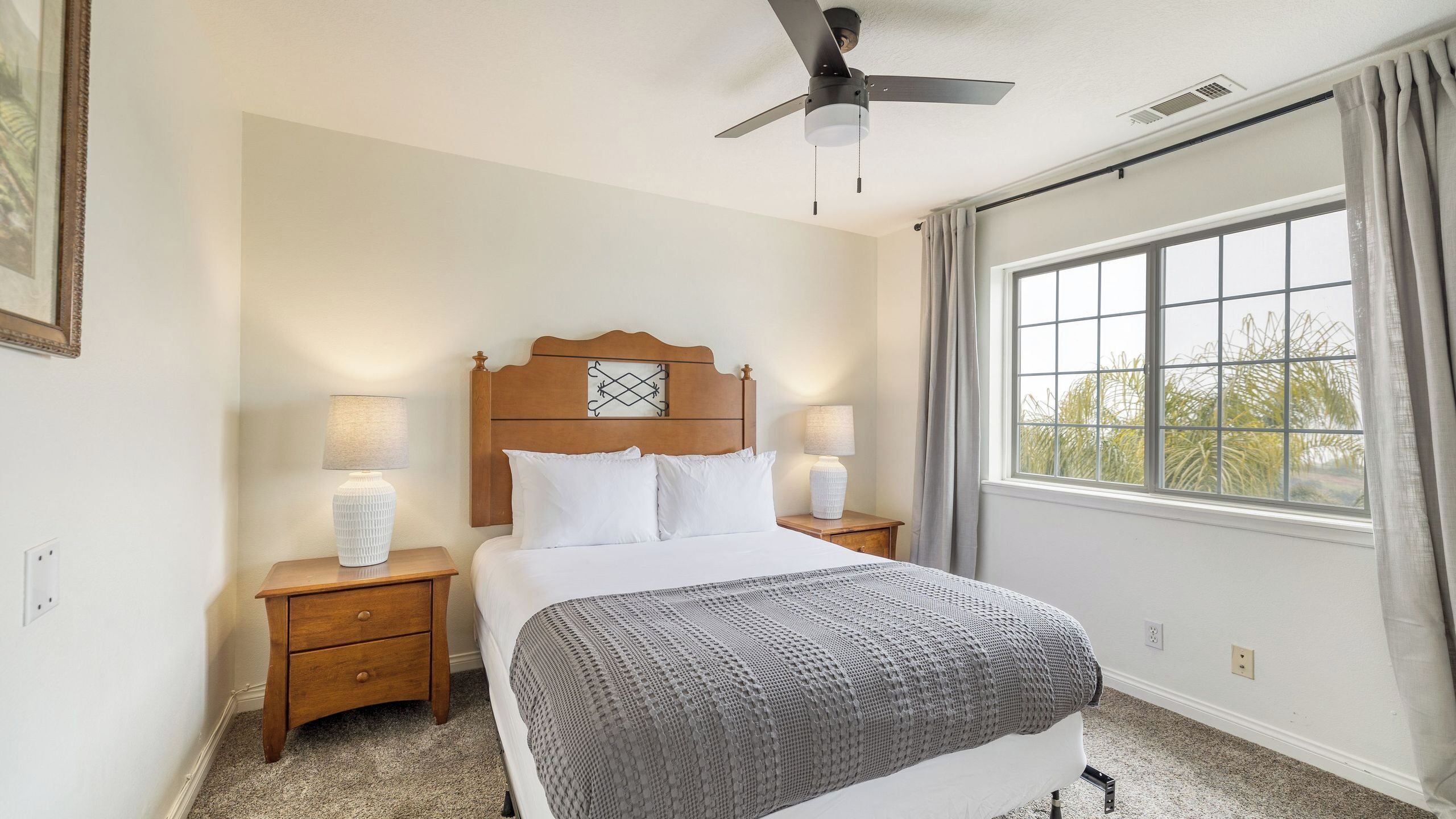 A bedroom with a bed, two bedside tables with lamps, a ceiling fan, a window with curtains, and a view of trees outside.