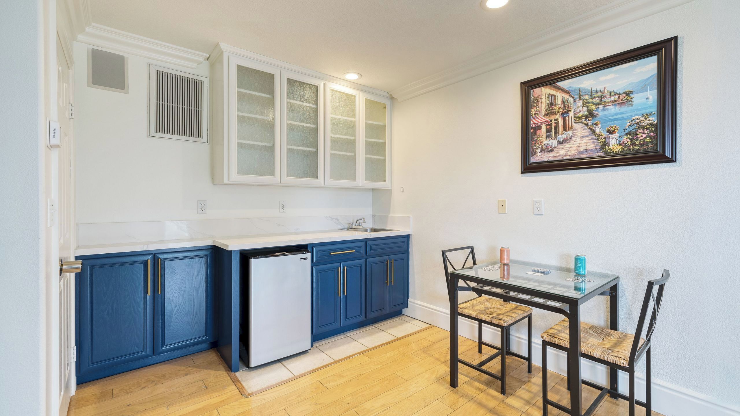 Una cocina compacta con refrigerador, armarios azules, una mesa de comedor con sillas y un cuadro en la pared.