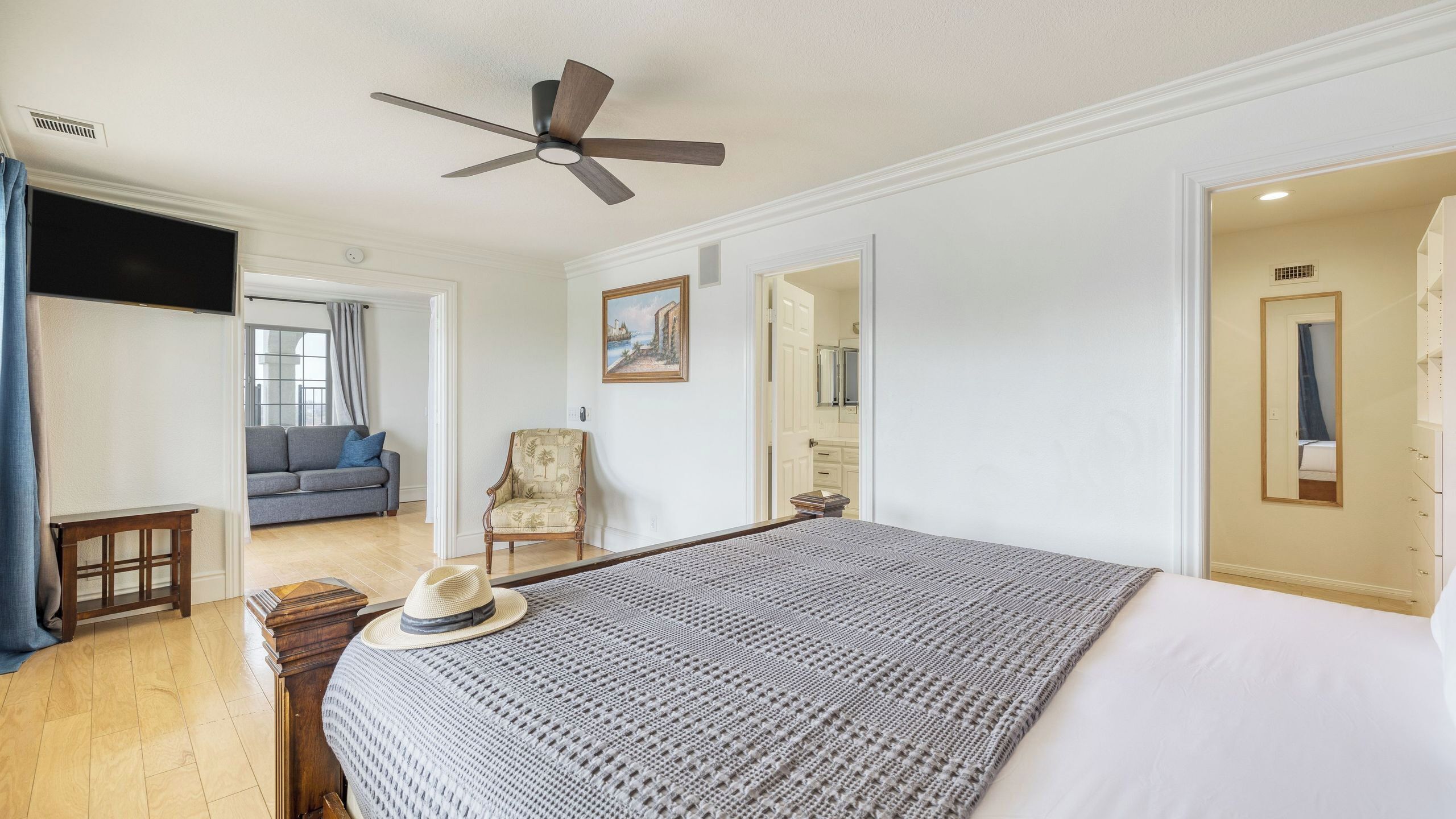 A bedroom with a bed, a ceiling fan, a chair, a sofa, and a painting on the wall.