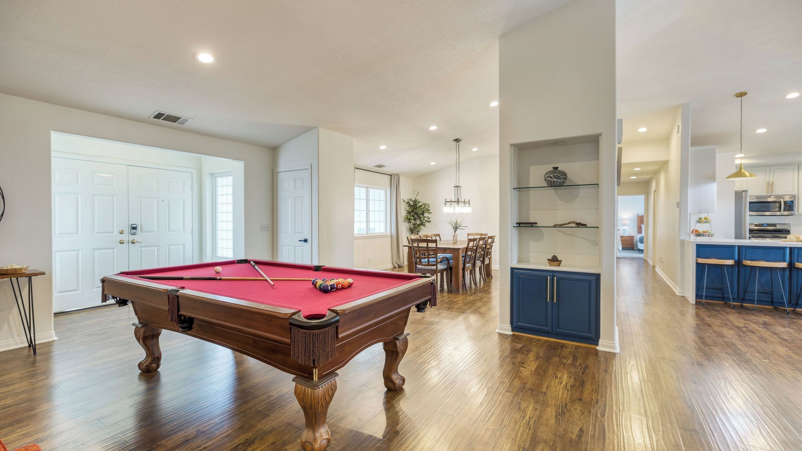 A spacious room with a pool table, wooden flooring, a dining area, and a kitchen with blue bar stools.