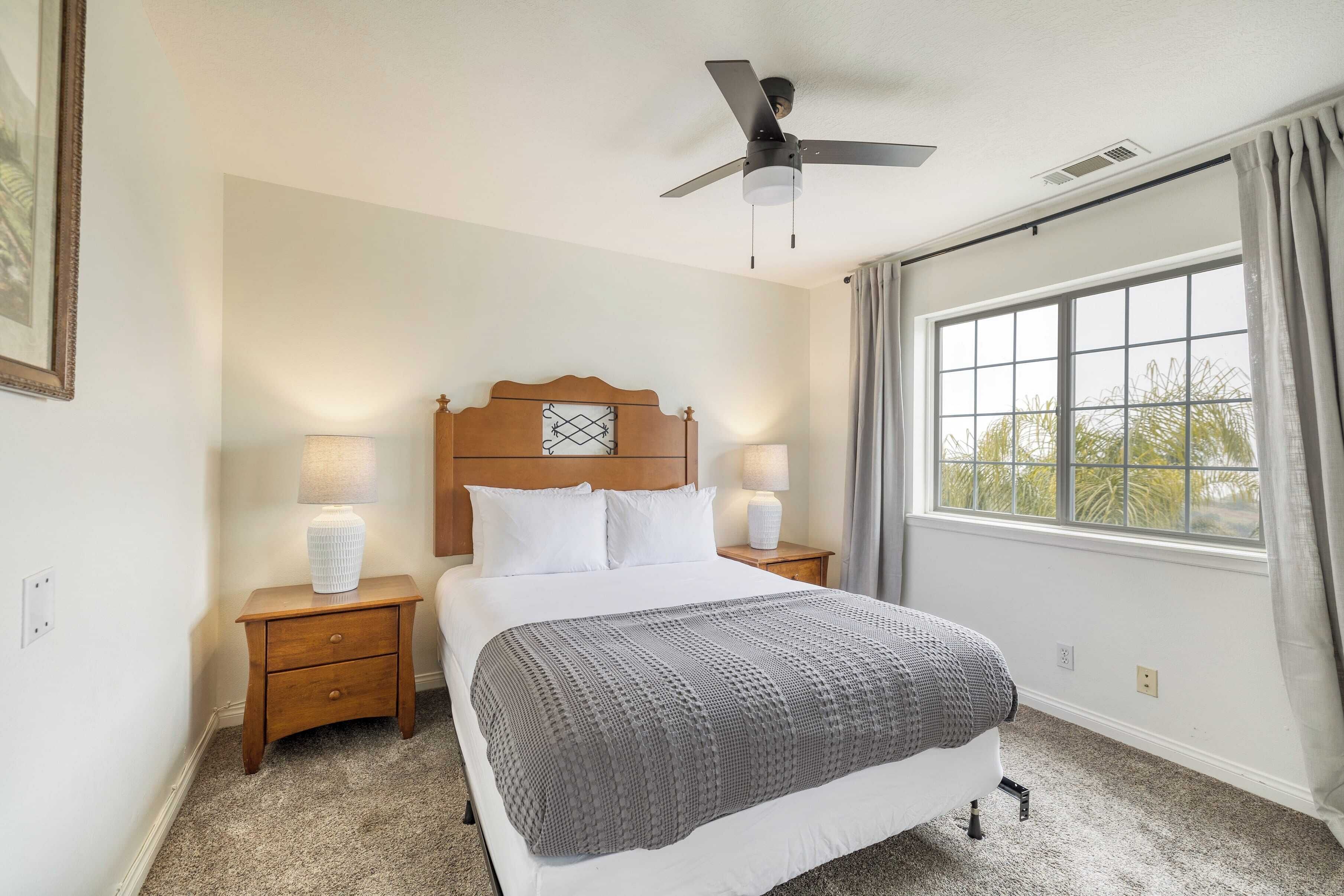 A bedroom with a bed, two bedside lamps, a ceiling fan, a window with curtains, and a framed picture on the wall.