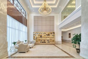 Un hall d’entrée spacieux avec un plafond haut, un lustre, un coin salon avec des canapés, une bibliothèque et une grande fenêtre.