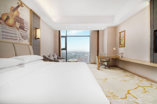 A hotel room with a bed, a desk, a chair, a small table, a large window, and a painting on the wall.