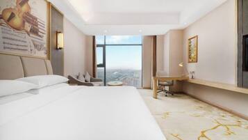 A hotel room with a bed, a desk, a chair, a small table, a large window, and a painting on the wall.