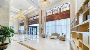 A spacious lobby with a high ceiling, large chandeliers, a grand staircase, and a seating area with sofas and a coffee table.