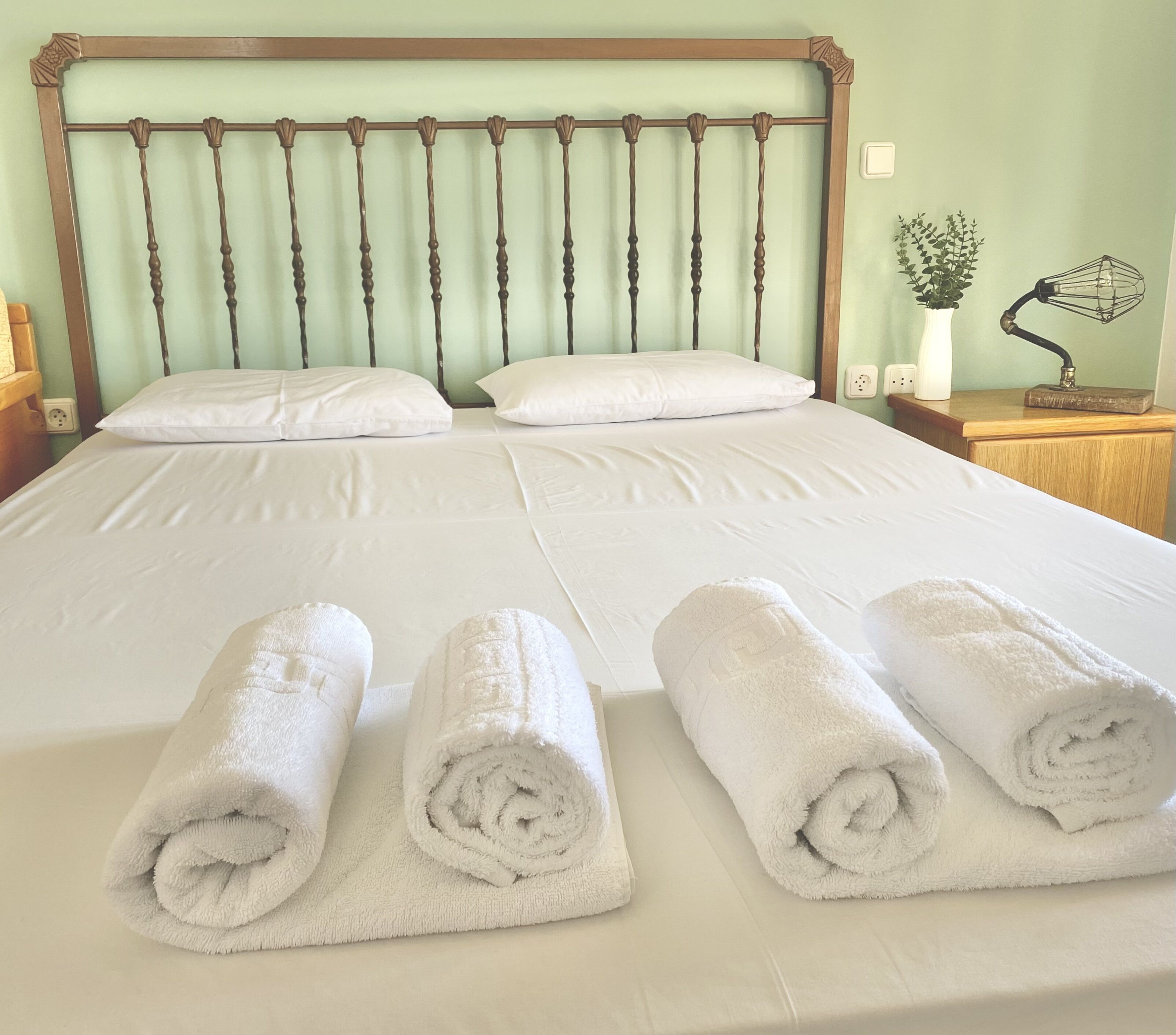 A neatly made bed with white linens and rolled towels, a wooden headboard, and a bedside table with a lamp and a plant.