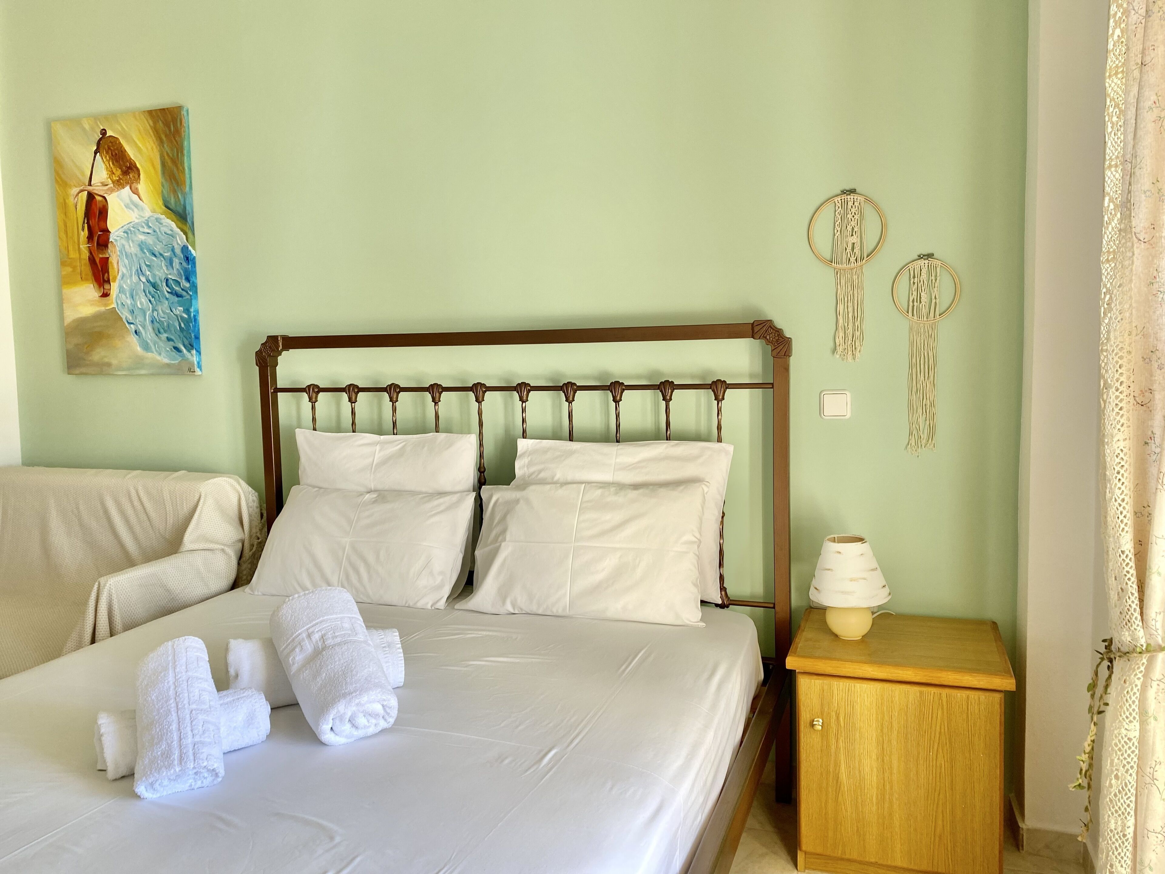 Cama bem arrumada com lençóis brancos e duas toalhas enroladas, mesa de cabeceira de madeira com abajur e pintura na parede de uma mulher tocando violoncelo.
