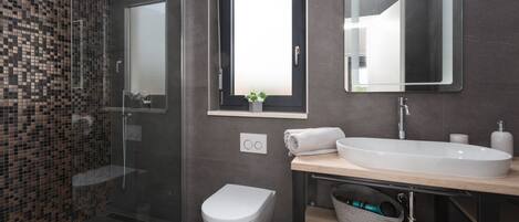 A modern bathroom with a glass shower enclosure, a toilet, a sink with a mirror, and a small window with a plant.