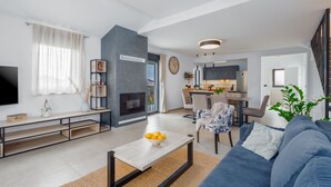 A modern living room with a sofa, coffee table, and a TV. There is a fireplace, a dining area, and a kitchen visible. - Villa for 4-6 people, pool, loungers, garden, panoramic sea view (Poreč)