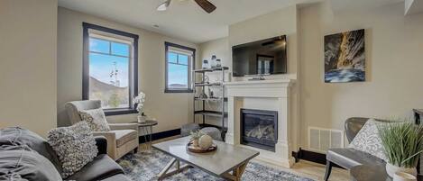 A living room with a fireplace, a ceiling fan, and a TV mounted above it.