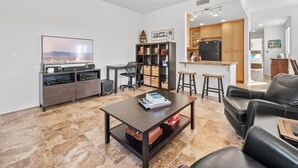 A modern living room with a flat-screen TV, a coffee table, a leather sofa, and a kitchen area with wooden cabinets and a refrigerator.