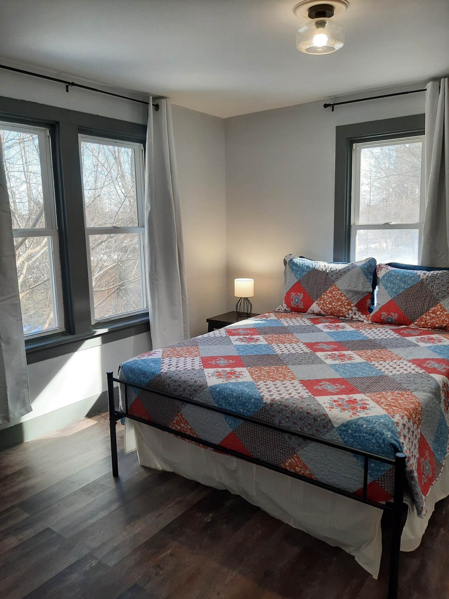 A bedroom with a bed, two windows, a nightstand, and a lamp.