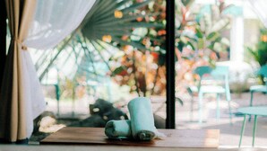 A room with a bed, a small table with a rolled-up towel, and a view of a garden through sheer curtains.