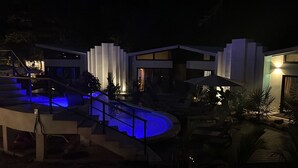A modern outdoor pool area with a blue-lit water feature, a staircase, and a well-lit building with large windows.