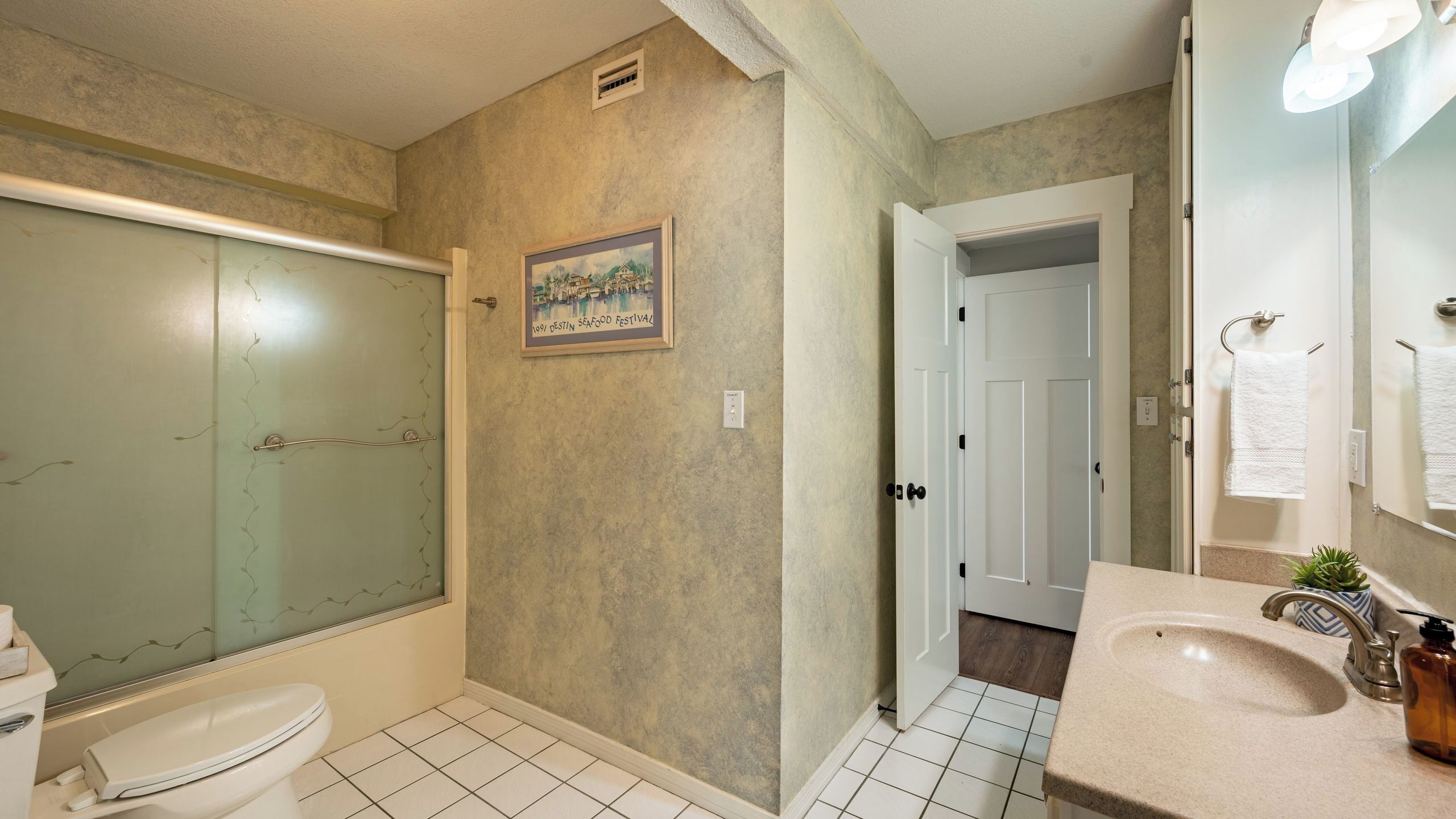A bathroom with a glass shower enclosure, a toilet, a sink with a mirror, and a door leading to another room.