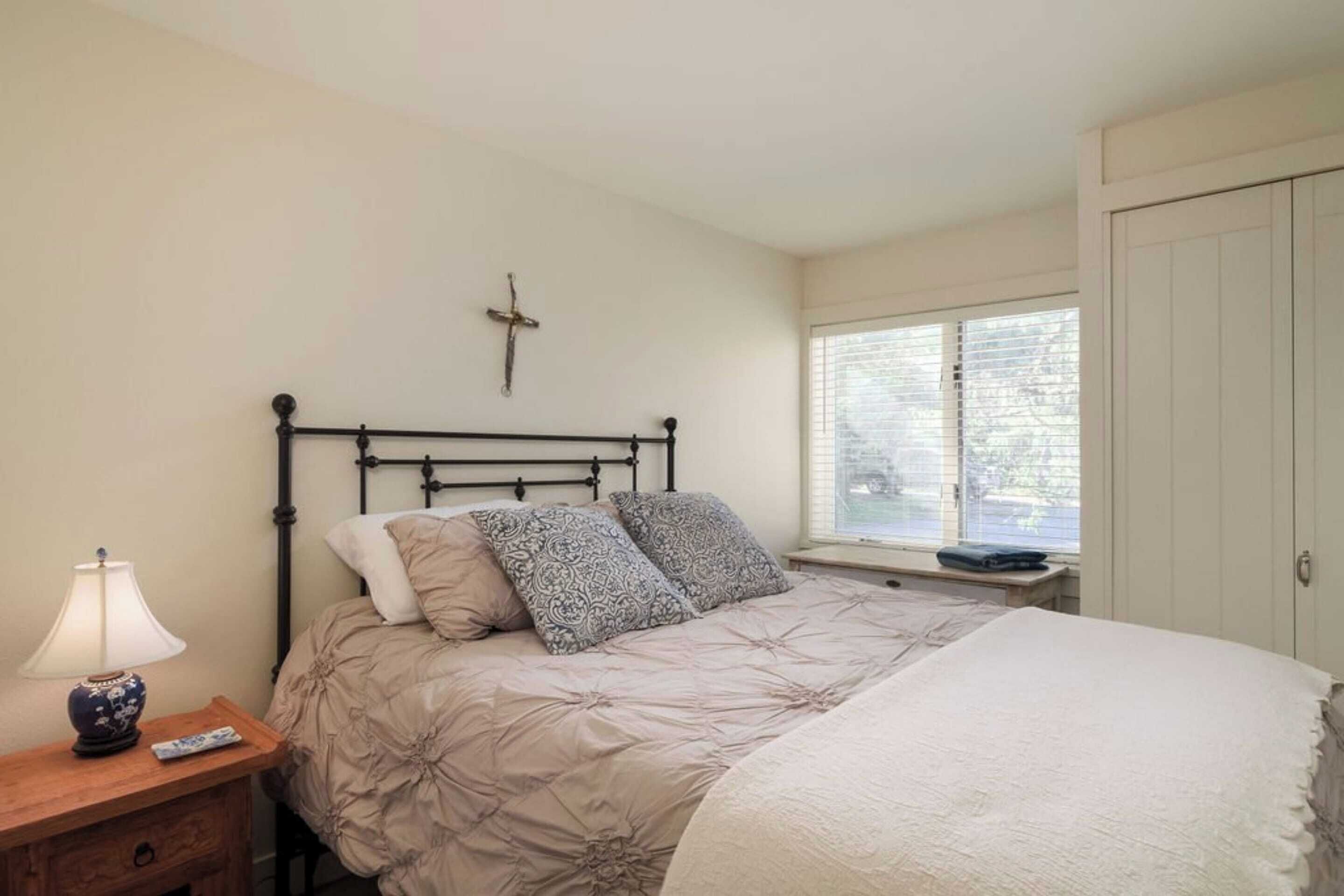 A bedroom with a bed, a nightstand, a lamp, a cross on the wall, and a window with blinds.