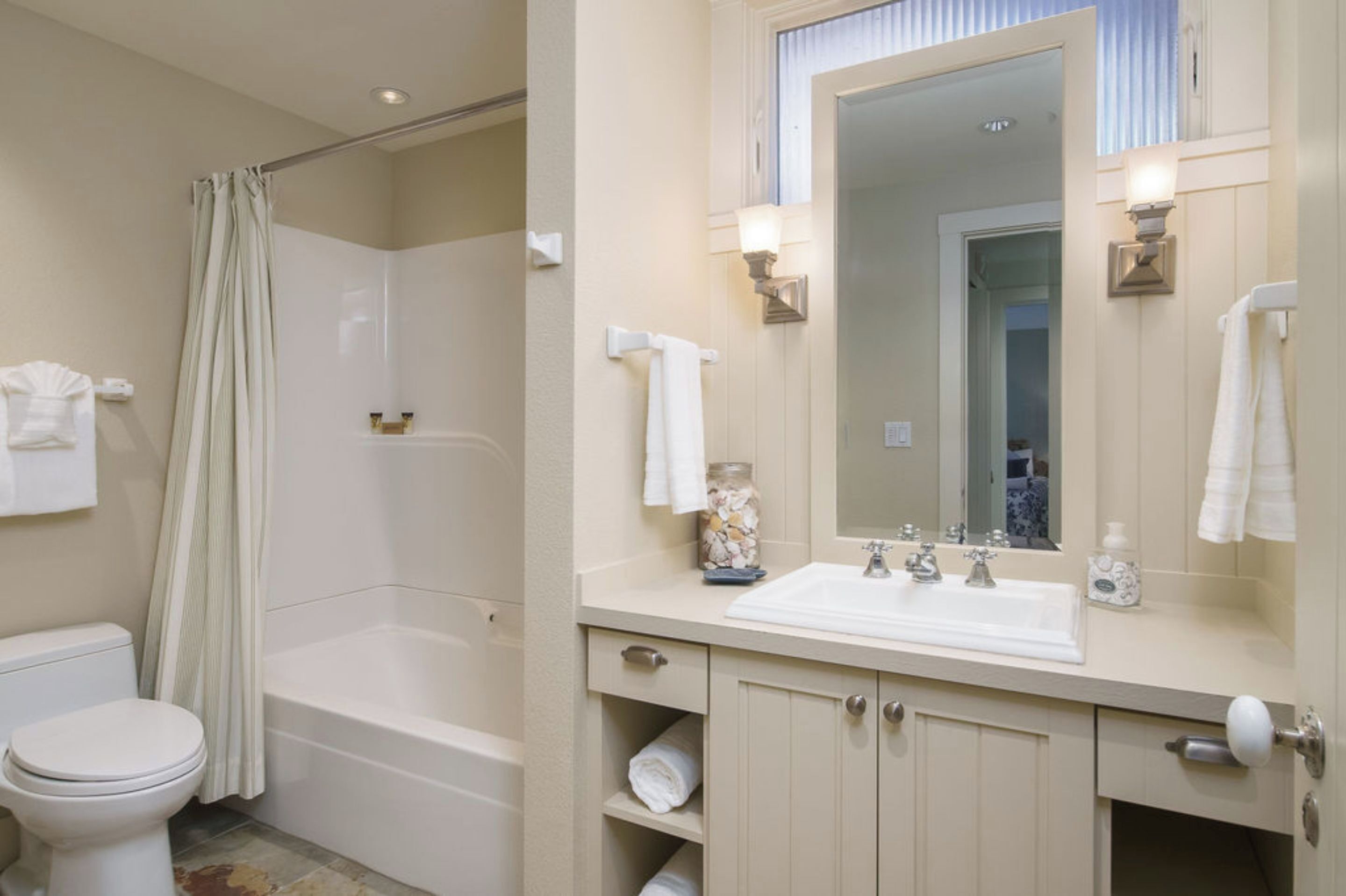 Een badkamer met toilet, badkuip, douchegordijn, wastafelmeubel met wastafel, spiegel, handdoekhouder en een decoratieve pot.