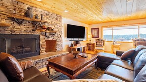 A cozy living room with a stone fireplace, wooden ceiling, leather sofas, and a large window offering a view.