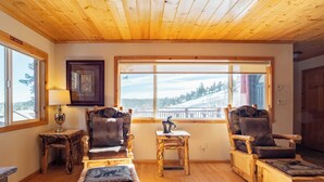 A cozy room with wooden flooring, two wooden chairs with cushions, a small table with a lamp, and a large window offering a view of a snowy landscape.