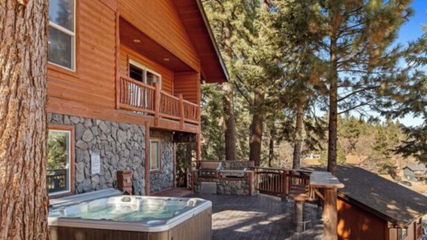 A wooden cabin with a hot tub, stone wall, and a balcony overlooking a forest.