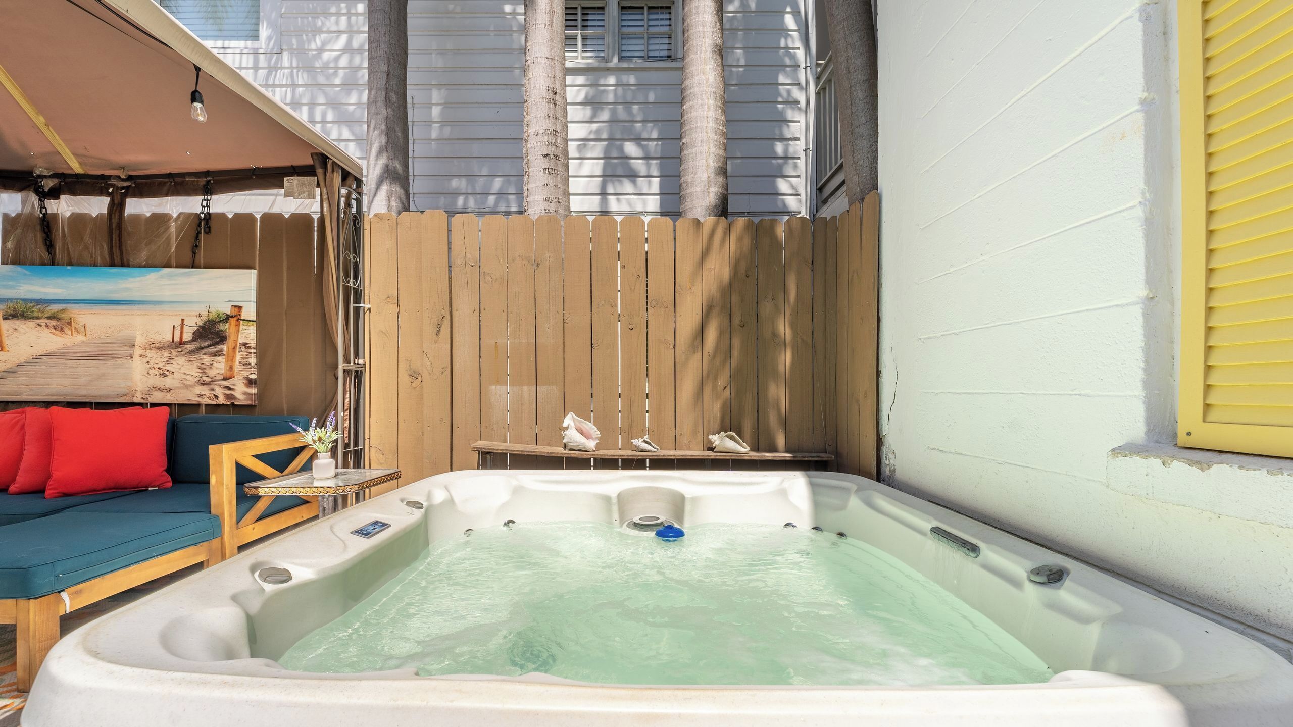 A hot tub in a backyard with a wooden fence, a beach scene painting, and a blue sofa.
