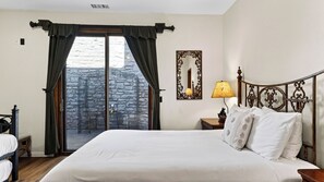A hotel room with a bed, a nightstand, a lamp, a mirror, and a view of a stone building through a window with curtains. - Walk to Lake 6BR Cabin w/ Game Room, Hot Tub Big Bear Castle (Big Bear Lake)