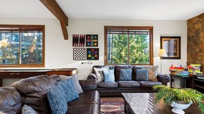 A living room with a brown leather sofa, a coffee table, and a pool table. There are windows with blinds, a lamp, and a plant. - Walk to Lake 6BR Cabin w/ Game Room, Hot Tub Big Bear Castle (Big Bear Lake)