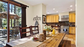A modern kitchen with wooden cabinets, a dining area with a table set for two, and a view of the outdoors through large windows. - Walk to Lake 6BR Cabin w/ Game Room, Hot Tub Big Bear Castle (Big Bear Lake)
