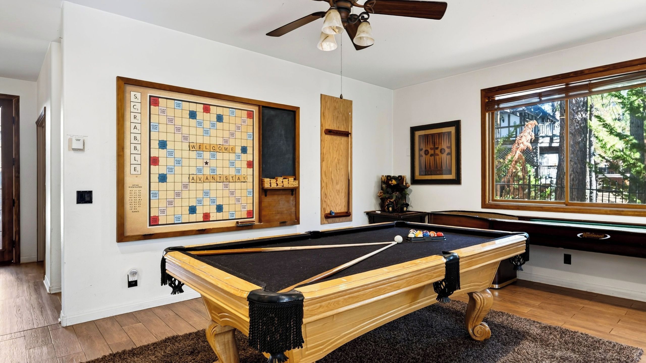 Uma mesa de bilhar com moldura de madeira e feltro preto, um jogo de Scrabble com a palavra 'WELCOME' e uma janela com vista para árvores e um edifício.