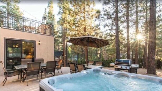A hot tub with a grill and chairs in a forested outdoor setting. - Walk to Lake 6BR Cabin w/ Game Room, Hot Tub Big Bear Castle (Big Bear Lake)