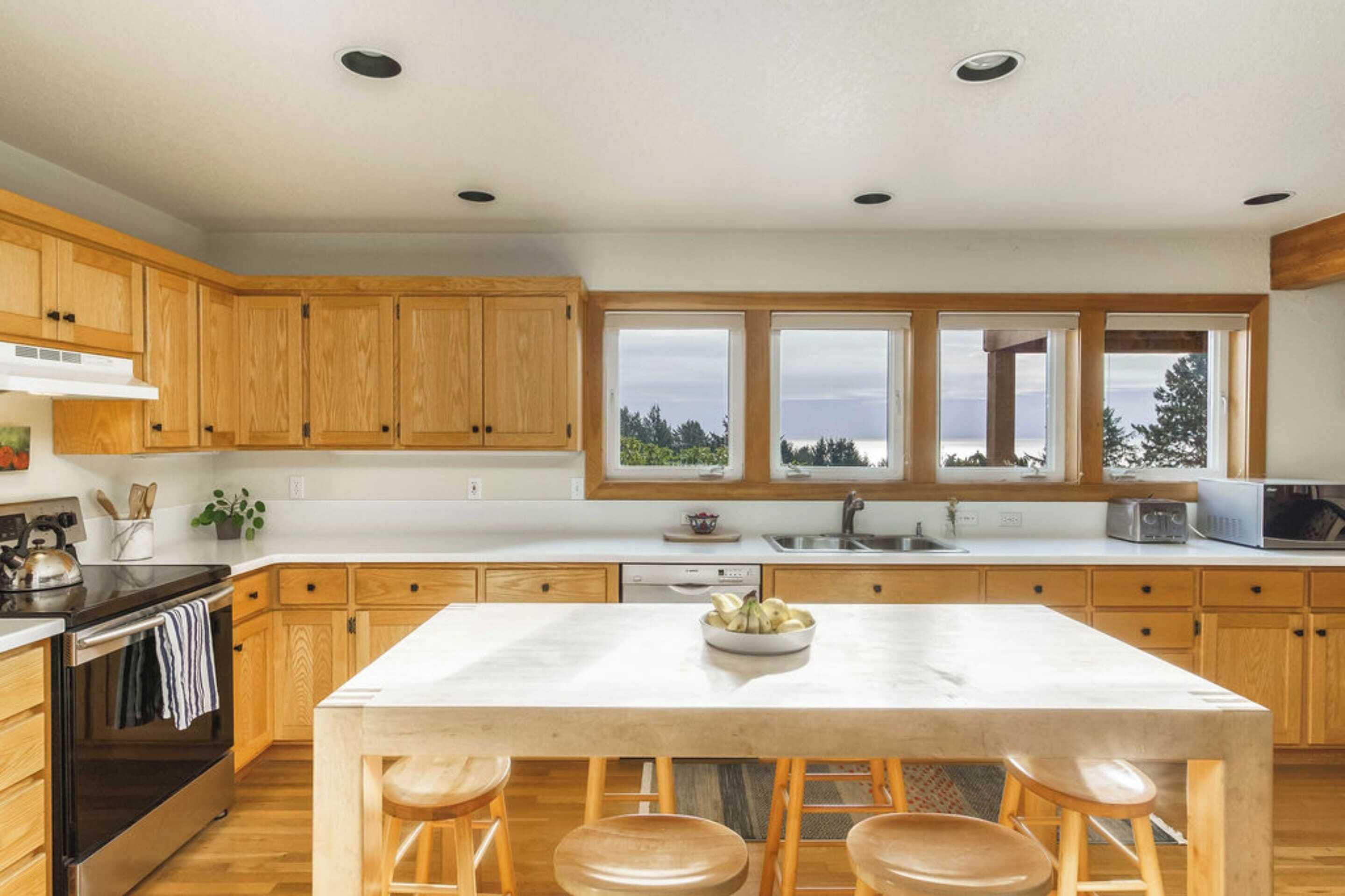 Een moderne keuken met houten kastjes, een centraal eiland met krukken, een ingebouwde oven en een groot raam met uitzicht op de omgeving.