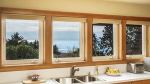 A kitchen with a large window overlooking a body of water, a sink, a toaster, and a bowl of fruit. - Coastal Getaway in Manzanita w Fire Pit, Guest House & Views Seagrove by Avantstay (Manzanita)
