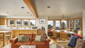 A spacious living area with a leather sofa, dining table, and chairs, wooden cabinets, and large windows.