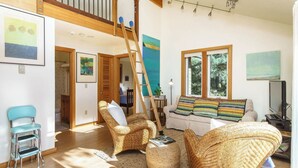 A spacious living room with a high ceiling, a wooden staircase, a sofa with colorful cushions, a television, and a painting on the wall. - Coastal Getaway in Manzanita w Fire Pit, Guest House & Views Seagrove by Avantstay (Manzanita)