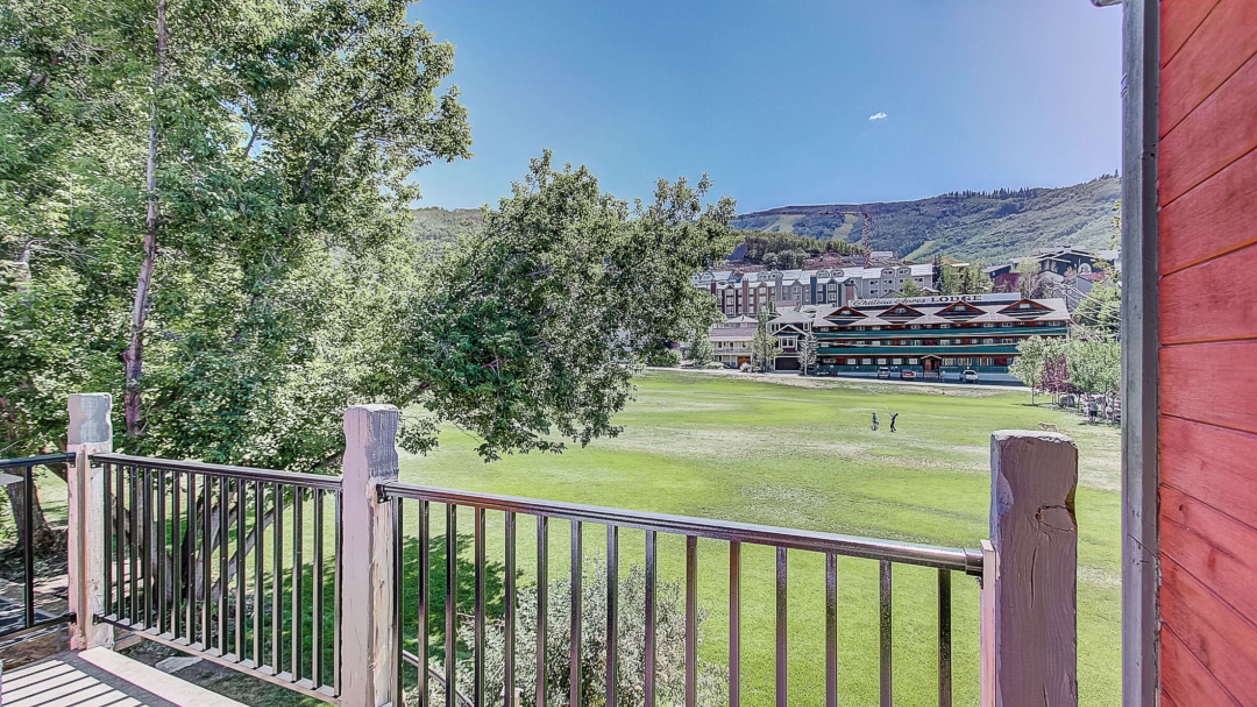 Une vue depuis un balcon doté d’une balustrade en bois, donnant sur une pelouse verdoyante, un bâtiment de style chalet et un paysage montagneux.