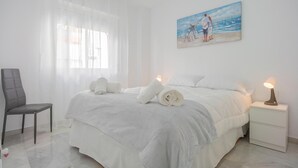 Una camera d'albergo con un letto, una sedia, un comodino e un quadro raffigurante una spiaggia.