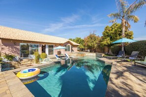 A swimming pool with lounge chairs and a sun umbrella, surrounded by a patio area with a tiled roof and a palm tree. - Luxury Oasis w/ Pool + Hot Tub, Fire Pit, Games & Big Yard Larrea by Avantstay (Indio)