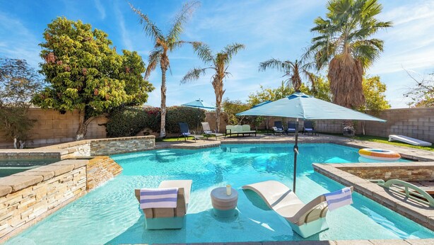 Une piscine entourée de chaises longues et de parasols, bordée de palmiers et d’un mur de pierre.