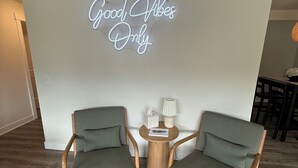 A room with two green upholstered chairs, a small wooden table with a lamp, and a neon sign on the wall that reads "Good Vibes Only". - Convenient modern 2br home, 5 mins from airport. (Tukwila)