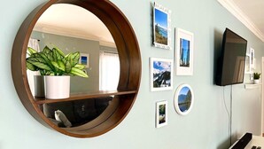 Une pièce avec un miroir rond, une plante en pot, des tableaux encadrés accrochés au mur, un téléviseur à écran plat et un meuble blanc.