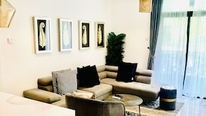 Ein modernes Wohnzimmer mit einer Couch, Sesseln, einem Couchtisch und einem Essbereich. An den Wänden befinden sich gerahmte Kunstwerke und eine große Pflanze.